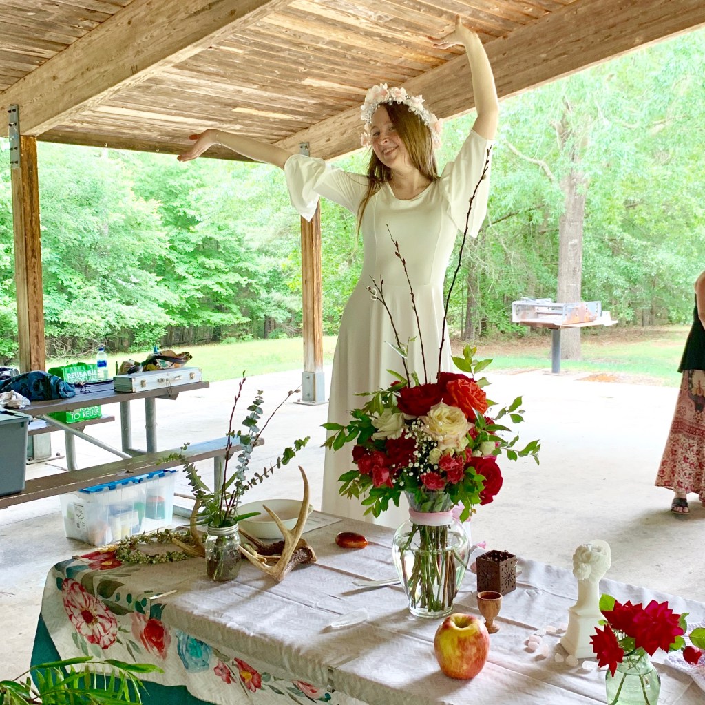 Stephanie wearing a white dress with bell sleeves and a flower crown, dancing on a picnic table during a Beltane ritual, behind an altar to Aphrodite and the Lord of the Wild Wood. On the altar in front to the right side is a marble statue of Aphrodite, roses in vases, an apple, rose quartz crystals. On the left side of the altar is a pair of deer antlers, a carnelian stone, and greenery.