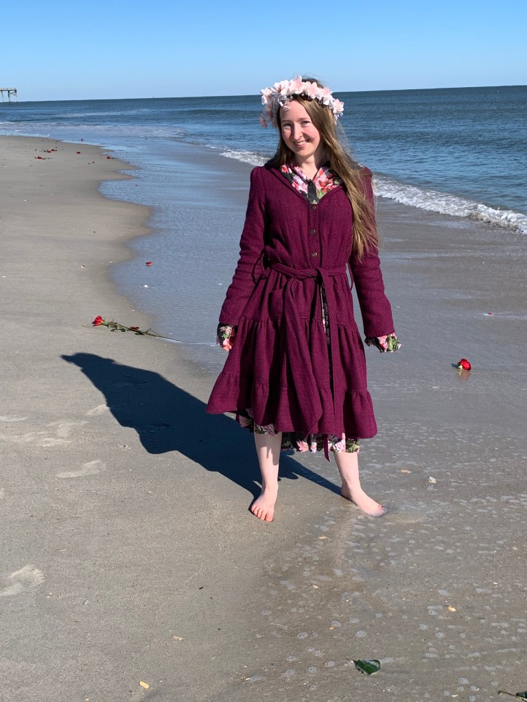 Stephanie crowned in pink and white flowers wearing a fuchsia jacket dress, standing barefoot on a beach scattered with roses as the waves crash to shore.