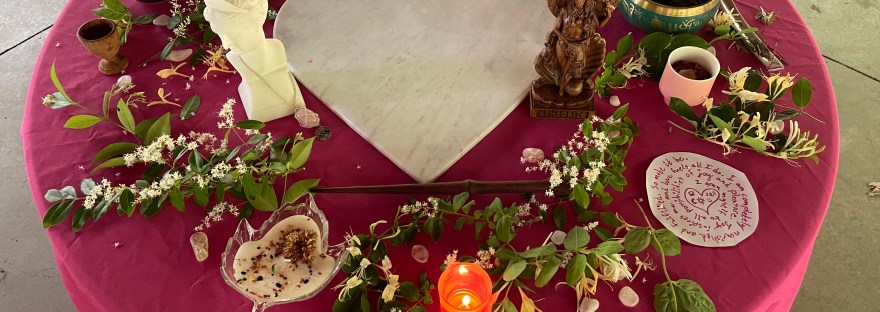 Photo of a Beltane altar with a white marble heart in the middle of a pink tablecloth, surrounded by statues of Aphrodite and greenery