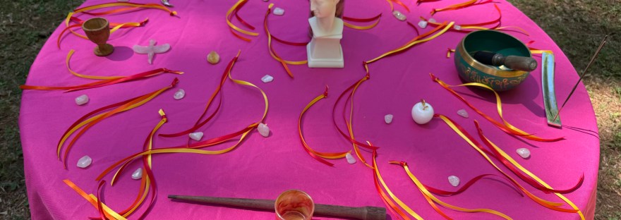 Beltane Altar on a pink tablecloth with a statue of Aphrodite at the center, surrounded by colorful bunches of ribbon